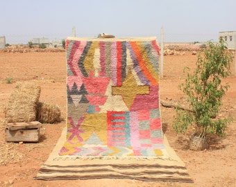 May include: A colorful, geometric patterned rug with a variety of colors, including pink, red, yellow, blue, and green. The rug is folded and laid out on a dirt surface.