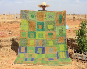May include: A colorful handwoven rug with a geometric pattern of squares in shades of green, brown, blue, and yellow. The rug is being held up by a person wearing a straw hat.