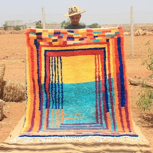 May include: A man holds up a colorful woven rug with a geometric pattern. The rug has a yellow, blue, and red color scheme. The rug is being held in front of a dirt background.