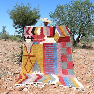 May include: A colorful Moroccan rug with geometric patterns in shades of red, yellow, blue, and white. The rug is woven with a variety of textures and is displayed outdoors in a desert landscape.
