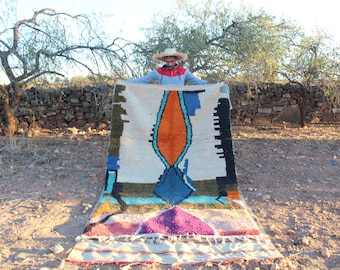 May include: A colorful Moroccan rug with geometric patterns in shades of blue, orange, green, and pink. The rug is being held up by a person wearing a straw hat.