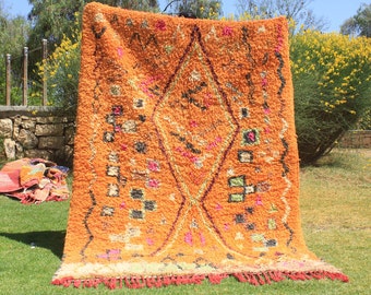 May include: A large, orange Moroccan rug with a geometric pattern. The rug has a fringe border and is made of wool.