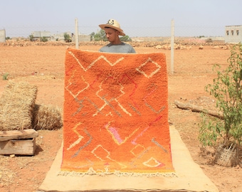 Op de afbeelding: Een handgeweven oranje kleed met een geometrisch patroon in tinten wit, roze en grijs. Het kleed wordt vastgehouden door een persoon met een strohoed.