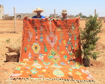May include: Two people hold up a large, colorful Moroccan rug with a bright orange background and intricate patterns in shades of pink, yellow, green, and blue. The rug has a fringe border.