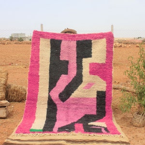 May include: A pink, black, and white Moroccan rug with a geometric pattern. The rug is hanging on a clothesline in a field.
