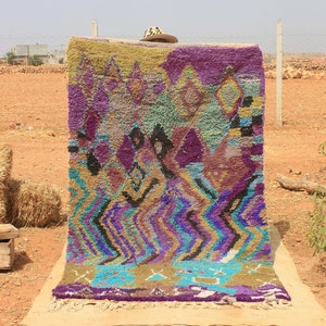 May include: A colorful Moroccan rug with a geometric pattern in shades of purple, blue, green, and gold. The rug is hanging on a wall in a desert landscape.