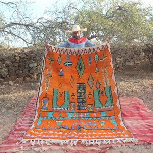 May include: A man holds up a large, orange and blue Moroccan rug with intricate geometric patterns. The rug has a fringe border and is woven with wool.