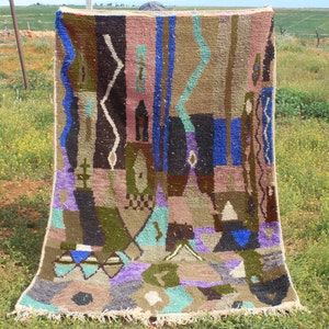 May include: A colorful abstract rug with geometric patterns in shades of brown, blue, green, purple, and pink. The rug has a fringe border and is displayed against a backdrop of green grass and wildflowers.