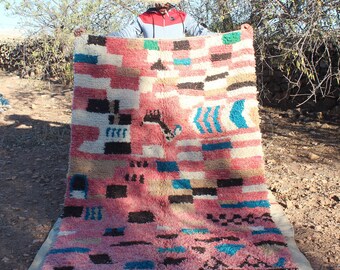 May include: A person holds up a large, colorful rug with a geometric pattern. The rug is made of wool and has a variety of colors, including pink, blue, black, and white.