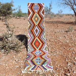 May include: A colorful Moroccan rug with a geometric diamond pattern in shades of red, orange, yellow, blue, green, and purple. The rug is woven with wool and has a fringe on both ends.