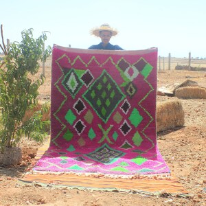 Pode incluir: Um tapete marroquino rosa e verde com um padrão geométrico. O tapete está sendo segurado por uma pessoa usando um chapéu de palha.