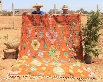 May include: Two people hold up a large, orange Moroccan rug with a colorful geometric pattern. The rug has a fringe border and is woven with wool.