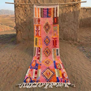 Peut inclure: Un tapis rectangulaire tissé à la main avec un fond rose et des motifs géométriques orange, bleu, jaune et rouge. Le tapis est suspendu à une tige en bois, avec une frange en bas. Le tapis est exposé à l'extérieur.