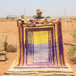 May include: A man holds up a colorful woven rug with a yellow, purple, and brown geometric pattern. The rug is being displayed outdoors on a dirt surface.