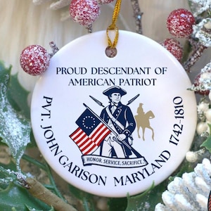 May include: A white ceramic ornament with a patriotic design. It features text that reads "PROUD DESCENDANT OF AMERICAN PATRIOT" and "PVT. JOHN GARISON MARYLAND 1742-1810." The ornament includes an illustration of a soldier and the American flag. A smaller emblem reads "DESIGNED AND MADE IN AMERICA."