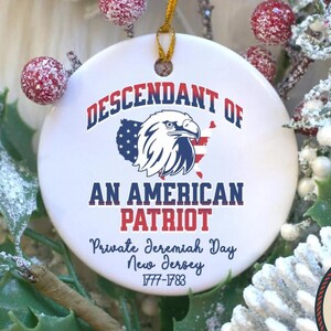 May include: A white ceramic ornament with the text "DESCENDANT OF AN AMERICAN PATRIOT" and an eagle design. The ornament also includes the text "Private Jeremiah Day, New Jersey, 1777-1783" and a "Designed and Made in America" seal. The ornament is surrounded by festive greenery.
