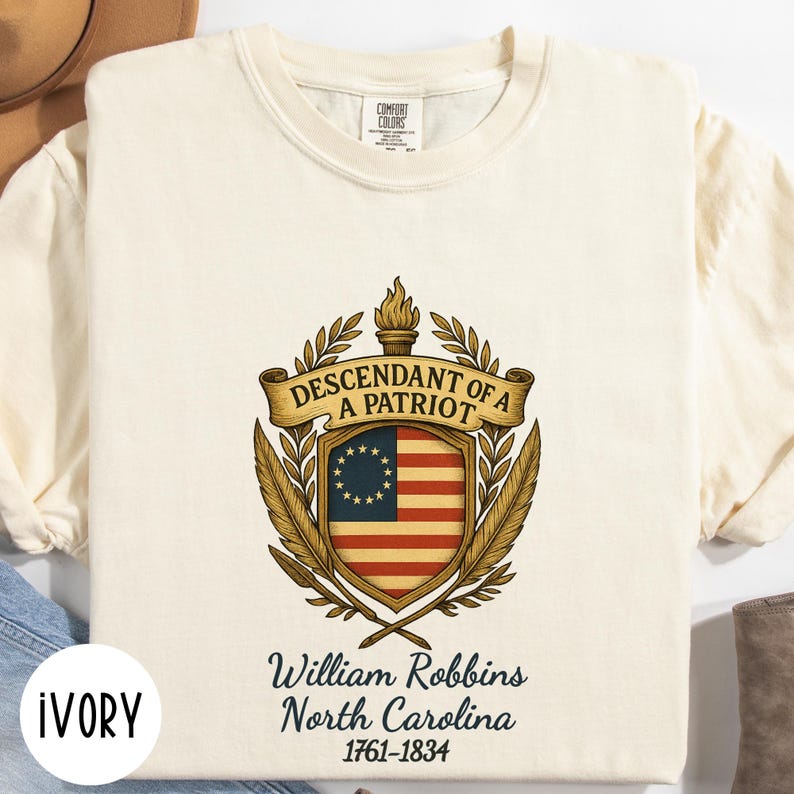 Op de afbeelding: Ivoor kleurig t-shirt met een patriottisch ontwerp. Het ontwerp toont een wapen met een banner met de tekst "Descendant of a Patriot" boven een vlag met sterren en strepen. Daaronder staat de tekst "William Robbins North Carolina 1761-1834."
