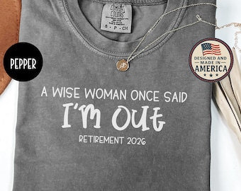 May include: Gray Comfort Colors t-shirt with the text "A Wise Woman Once Said I'm Out Retirement 2026." The shirt has a crew neck and short sleeves. A silver necklace with a pendant is draped across the shirt. The shirt is designed and made in America.