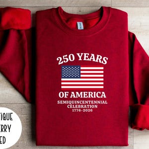 Puede incluir: Sudadera rojo cereza antiguo con el texto "250 YEARS OF AMERICA" y una bandera estadounidense. La sudadera celebra la Semiquincentennial Celebration de 1776-2026.