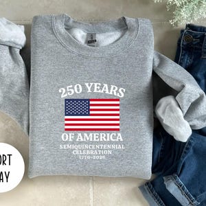 Puede incluir: Una sudadera gris con el texto "250 YEARS OF AMERICA" y una bandera estadounidense. La sudadera tambi&eacute;n incluye el texto "Semiquincentennial Celebration 1776-2026". La sudadera se muestra con unos vaqueros azules.
