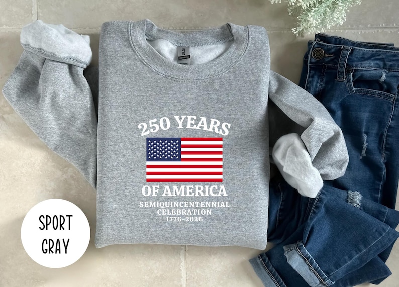 Puede incluir: Una sudadera gris con el texto "250 YEARS OF AMERICA" y una bandera estadounidense. La sudadera tambi&eacute;n incluye el texto "Semiquincentennial Celebration 1776-2026". La sudadera se muestra con unos vaqueros azules.