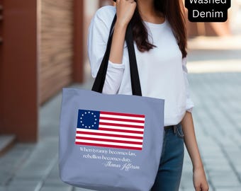 Op de afbeelding: Een lichtblauwe tote bag met het Amerikaanse Betsy Ross vlagontwerp. De tas heeft zwarte bandjes en bevat de quote "When tyranny becomes law, rebellion becomes duty." van Thomas Jefferson.