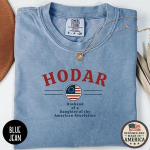 May include: A light blue t-shirt with the word "HODAR" in red block letters above a graphic of the American flag. Below the flag graphic, the text reads "Husband of a Daughter of the American Revolution." A gold necklace rests on the shirt.