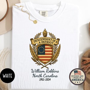 Op de afbeelding: Witte sweatshirt met een patriottisch ontwerp. De afbeelding bevat een schild met een koloniale Amerikaanse vlag, de tekst "DESCENDANT OF A PATRIOT" en de naam "William Robbins, North Carolina 1761-1834". De sweatshirt heeft de tekst "WHITE" linksonder.