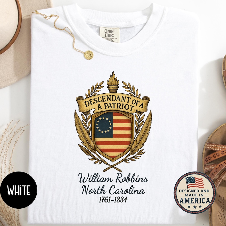 Op de afbeelding: Witte sweatshirt met een patriottisch ontwerp. De afbeelding bevat een schild met een koloniale Amerikaanse vlag, de tekst "DESCENDANT OF A PATRIOT" en de naam "William Robbins, North Carolina 1761-1834". De sweatshirt heeft de tekst "WHITE" linksonder.