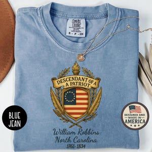 Op de afbeelding: Lichtblauw Comfort Colors T-shirt met een patriottisch ontwerp. Het ontwerp bevat de tekst "DESCENDANT OF A PATRIOT" boven een schild met de Amerikaanse vlag en de tekst "William Robbins North Carolina 1761-1834".