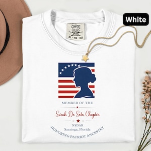 May include: White t-shirt with a patriotic design featuring a silhouette, stars, and stripes. The shirt includes text that reads "Member of the Sarah De Soto Chapter NSDAR Saratoga, Florida Honoring Patriot Ancestry."