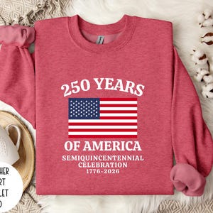 Puede incluir: Una sudadera roja escarlata con el texto "250 YEARS OF AMERICA" y una bandera estadounidense. La sudadera celebra el semiquincentenario de 1776-2026. La sudadera es de estilo deportivo jaspeado.