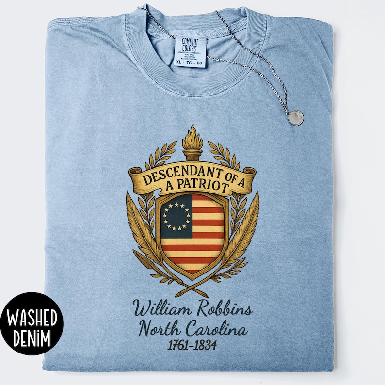 Op de afbeelding: Lichtblauwe gewassen denim t-shirt met een patriottisch wapenontwerp. Het wapen toont een vlag, een fakkel en de tekst "Descendant of a Patriot". Daaronder staat "William Robbins, North Carolina, 1761-1834."