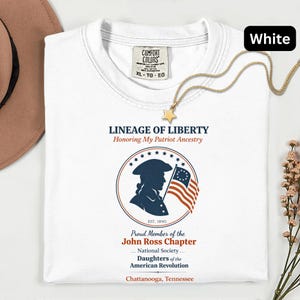 May include: White t-shirt with the text "LINEAGE OF LIBERTY" and a patriotic design featuring a silhouette and flag. The shirt also includes text for the John Ross Chapter of the Daughters of the American Revolution.