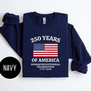 Puede incluir: Sudadera azul marino de manga larga con el texto "250 YEARS OF AMERICA" y una bandera estadounidense. La sudadera celebra el semiquincentenario de 1776-2026. Un c&iacute;rculo negro con la palabra "NAVY" est&aacute; en la esquina inferior izquierda.