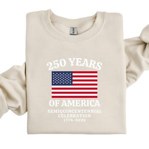 Puede incluir: Una sudadera color crema con el texto "250 YEARS OF AMERICA" sobre una bandera estadounidense. Debajo de la bandera, se lee "SEMIQUINCENTENNIAL CELEBRATION 1776-2026". La parte superior de manga larga est&aacute; doblada.