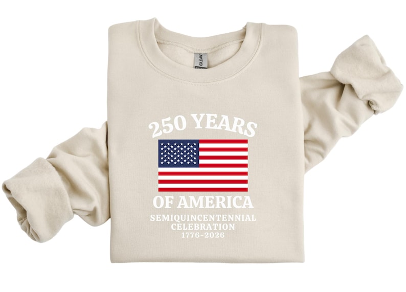 Puede incluir: Una sudadera color crema con el texto "250 YEARS OF AMERICA" sobre una bandera estadounidense. Debajo de la bandera, se lee "SEMIQUINCENTENNIAL CELEBRATION 1776-2026". La parte superior de manga larga est&aacute; doblada.