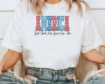 May include: White t-shirt with the word "AMERICA" in red and blue, outlined in white, with a dotted pattern. Below the word is the phrase "God Shed His Grace On Thee" in black. The shirt is paired with blue jeans.