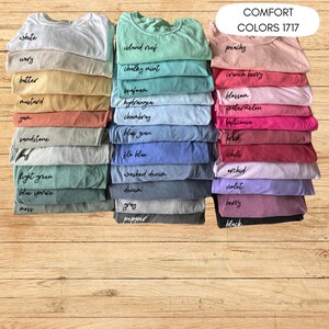 Op de afbeelding: Stapels opgevouwen t-shirts in verschillende kleuren, waaronder wit, geel, groen en roze. Elk shirt is gelabeld met zijn kleurnaam, zoals "peachy" en "blue jean". De tekst "COMFORT COLORS 1717" is ook zichtbaar.