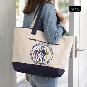 May include: A canvas tote bag with navy blue accents and straps. The bag features a circular emblem with the text "American Revolutionary Soldier" and an illustration of a soldier, flags, and a horse. The bag has a zippered pocket.