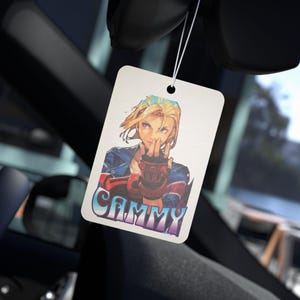 Puede incluir: Un ambientador de coche blanco con una ilustración colorida de una mujer con cabello rubio y un guante rojo. El texto "CAMMY" está impreso en un degradado azul y morado.