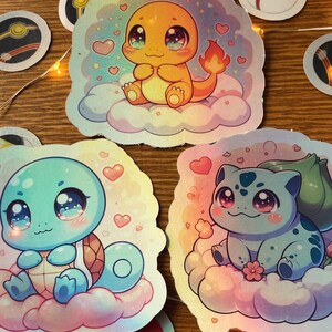 May include: Three holographic stickers featuring cartoon characters from the Pokemon franchise. The stickers feature Squirtle, Bulbasaur, and Charmander, each with large eyes and a pink blush. The characters are sitting on clouds with hearts and stars surrounding them.