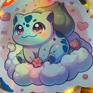 May include: A holographic sticker featuring a cartoon blue and green Pokemon character with large eyes, sitting on a cloud with pink hearts and flowers. The sticker has a shiny, iridescent finish.