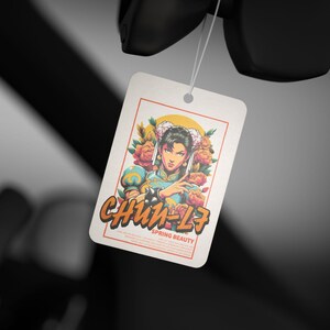 May include: A white car air freshener with a colorful illustration of a woman with flowers in her hair. The text "Chun-Li" and "Spring Beauty" is printed on the air freshener.
