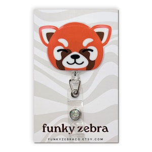 May include: Red panda cartoon face badge reel with a clear plastic retractable cord and a sparkly silver charm. The badge reel is on a white background with a zebra stripe pattern. The text "funky zebra" and "FUNKYZEBRACO.ETSY.COM" is printed on the background.