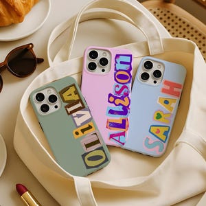 May include: Three phone cases with personalized names inside a white tote bag. The cases are in pastel colors: green, pink, and blue. The names "Olivia", "Allison", and "Sarah" are written in colorful, blocky letters. A croissant, sunglasses, and lipstick are also visible.