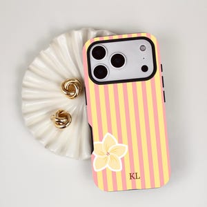 May include: A smartphone case featuring vertical yellow and pink stripes, a single light yellow plumeria flower graphic, and the black initials KL printed near the bottom. The case has a black camera cutout frame and a black border.