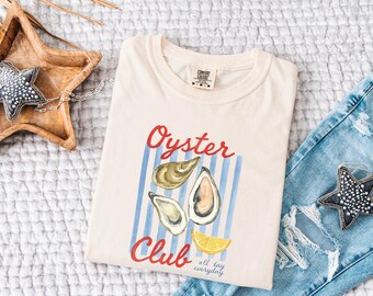 May include: Cream-colored t-shirt with the words "Oyster Club" in red script, featuring an oyster illustration and blue stripes. The shirt also has the words "all day everyday" in small black text.