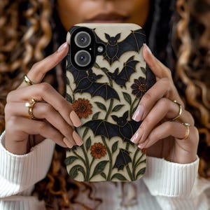 May include: A phone case featuring a repeating pattern of black bats, orange flowers, and green foliage against a cream background. The case has a black camera lens cutout and is held by a person.