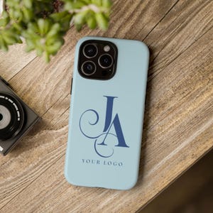 May include: A light blue phone case with a dark blue monogram of the letters "JA" and the text "YOUR LOGO". The phone case is on a wooden surface, with a camera and plant in the background.